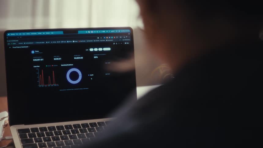 A woman is sitting at a desk with a laptop waiting for a graph to load on the screen