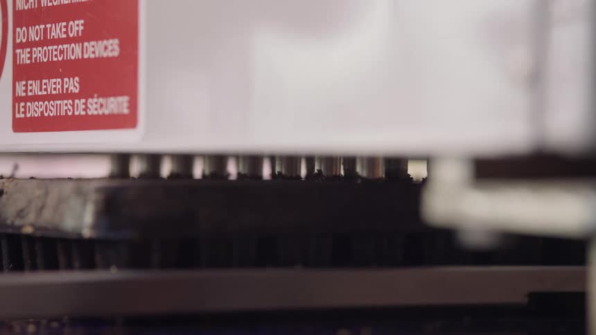 This is a closeup shot of an industrial machine possibly a highspeed printer conveyor or cutting tool in operationthe foreground shows a white panel with multilingual warning labels do not take off the protection devices while a black toothe