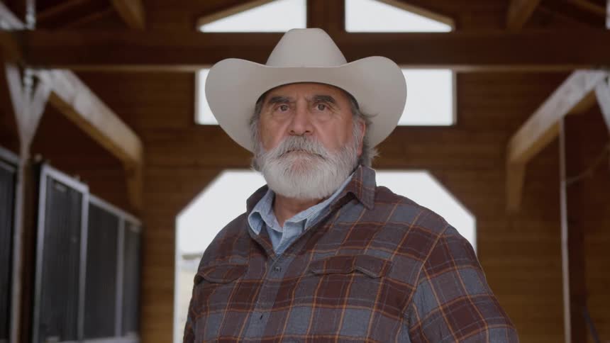 An elderly man wearing a cowboy hat is standing inside a barn