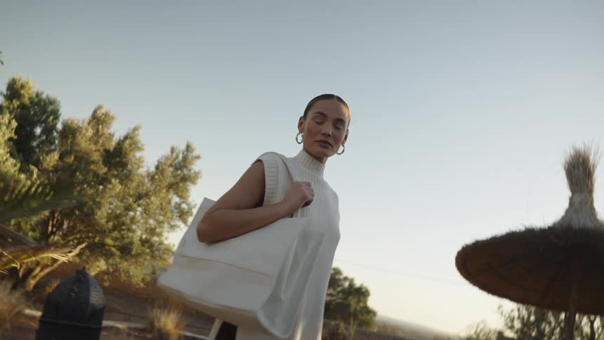 A young woman dressed in a stylish sleeveless white top and dark trousers is filmed from a low angle against a sunny slightly dusty landscape with palm trees and a straw umbrella visible she is carrying a large white tote bag over her shoulder
