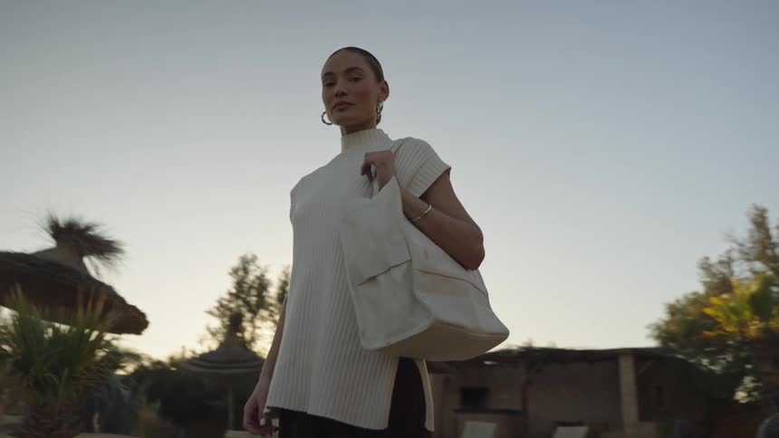 A young woman stands in an open space at midday holding a handbag wearing round large earrings and bangles