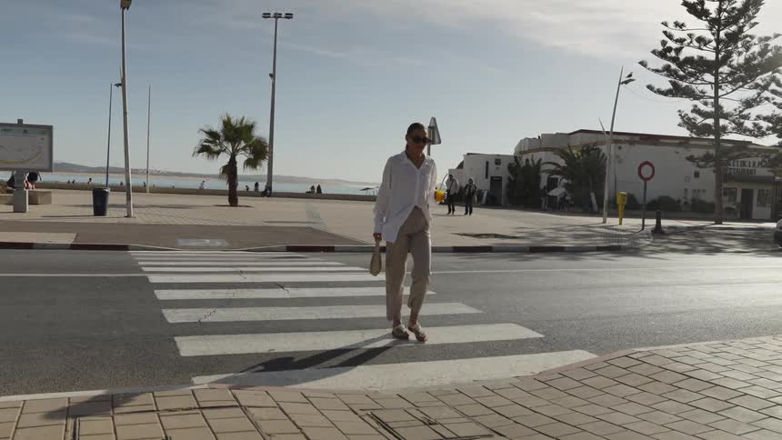 A woman in a white shirt and tan pants crosses a bright street near a palmlined promenade by the ocean