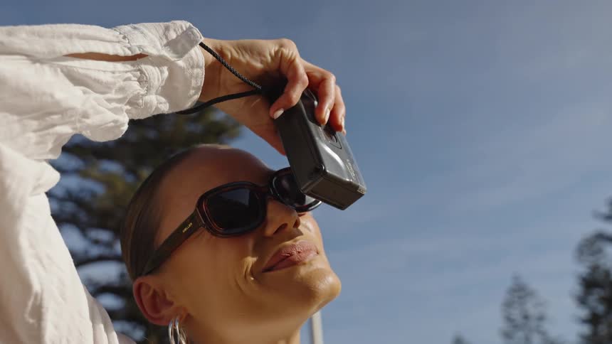 A woman wearing large sunglasses and a white blouse holds a small camera high above her head to capture a photo against a bright blue sky