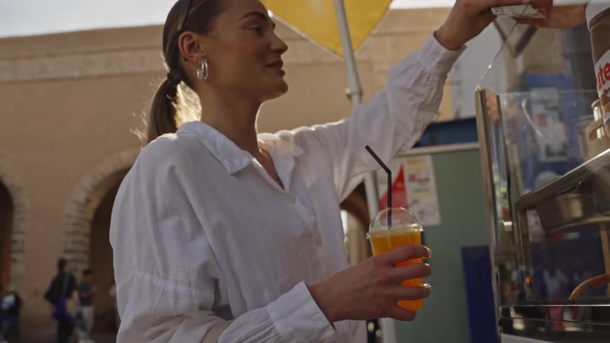 This clip shows a woman in a white shirt and sunglasses buying an orange drink from a street vendorshe is standing near a large archway in a busy outdoor area and is shown paying with cash