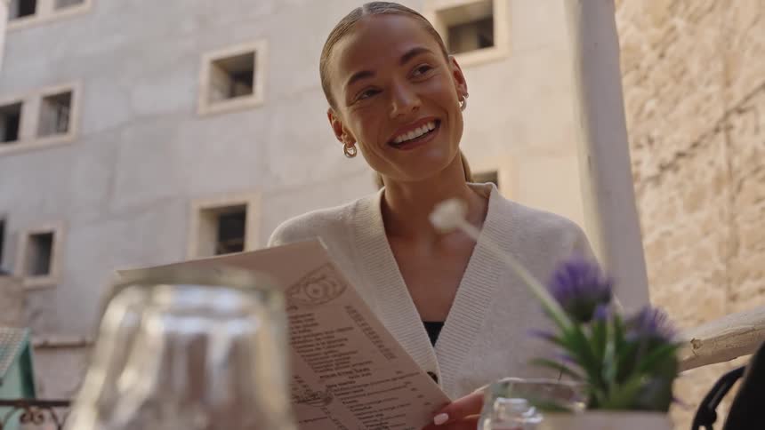 Young woman sitting on a table outdoors at a shop reading a menu and laughing during day time