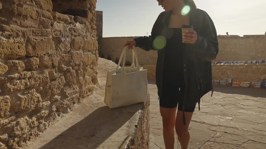 A woman in sunglasses walks along the stone path of an ancient fortress wall carrying a cup of coffee and a tote bag before pausing and sitting down on the stone ledge to rest