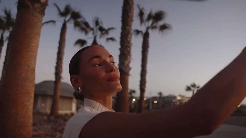 A smiling woman takes a selfie among palm trees at dusk