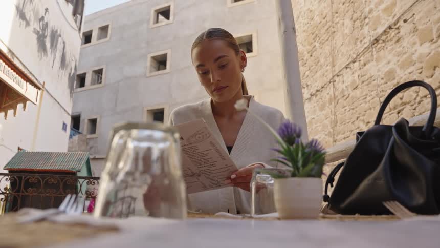 A young woman with her hair pulled back wearing a white cardigan sitting at an outdoor table and intently reading a menu with inverted glasses cutlery and a small potted plant against a backdrop of old lightcolored stone buildings