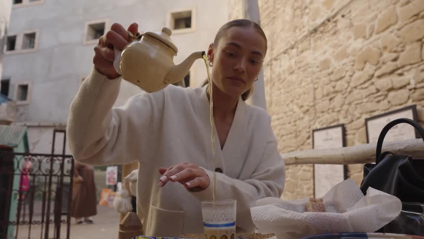 A woman pours amber tea from a white teapot into a small glass at an outdoor cafe