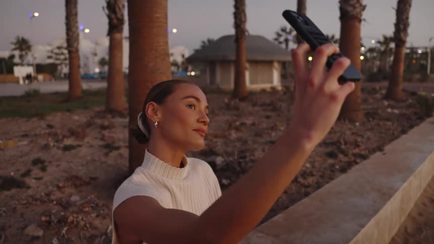 A woman is seat outside on a cement bench is talking a selfie near many palm trees in town