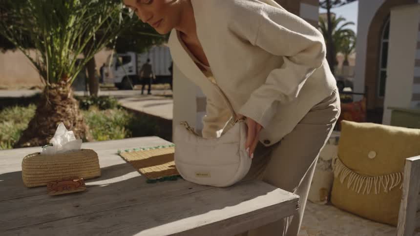 A woman wearing a lightcolored cardigan and trousers as she sits down at a table on a sunlit outdoor patio setting her white handbag down and then smiling as she settles into the moment
