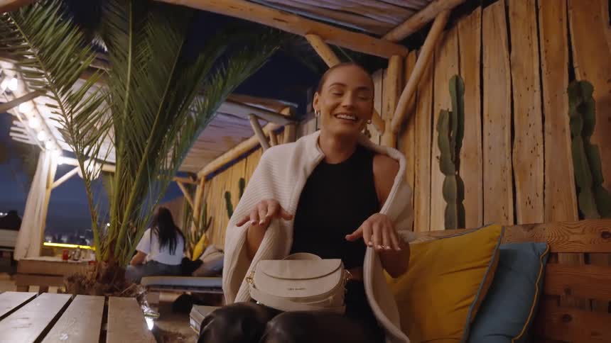 A woman wearing black clothes a white shawl and a small white handbag sits on a wooden bench in an outdoor rustic bar or lounge at night she looks up thoughtfully then turns to the camera smiling and laughing in the dimly lit tropical setting
