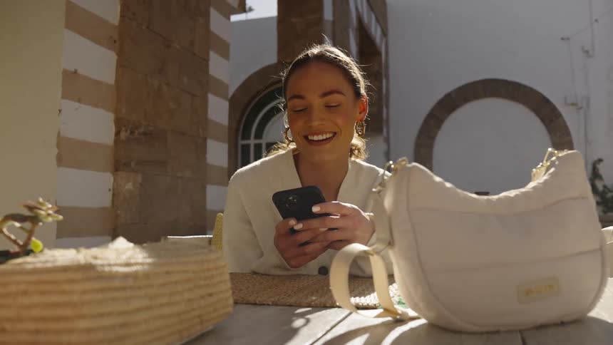 Woman sitting outside at a table smiles and laughs as she holds her phone