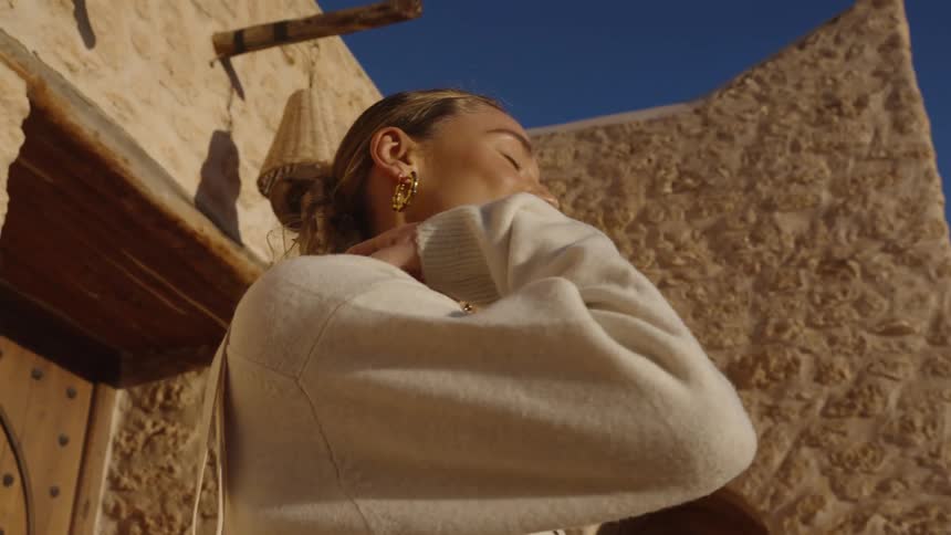 A young woman wearing a milkcolored sweatshirt stands in front of a building on a sunny day carrying a handbag and adjusting its strap on her shoulder