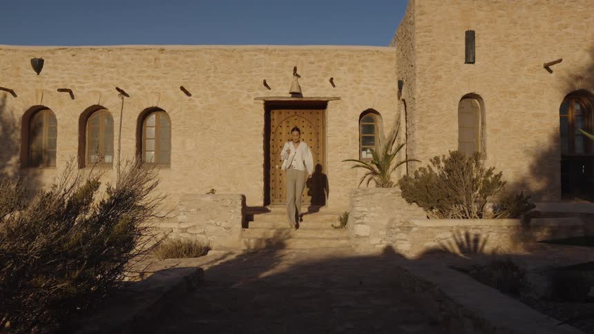A close view shows a woman in a light colored cardigan walking down steps from a wooden doorway set in a traditional stone villa in a desert environment