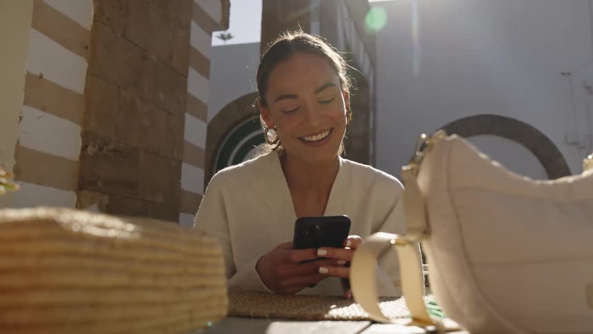 Woman sitting outside at a table smiles and laughs as she holds her phone