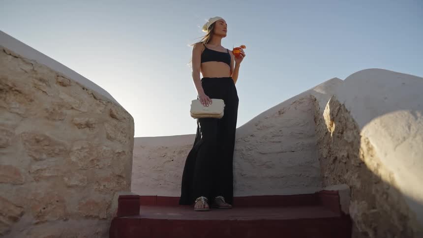 A woman stands on stone steps holding a cocktail and a bag on a sunlit terrace