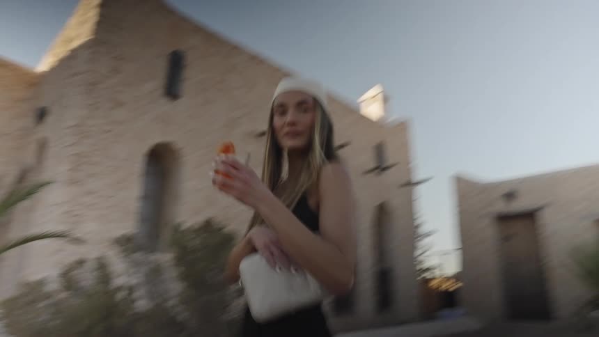 This dynamic clip shows a young woman holding an orange drink and a white bag walking or spinning quickly towards the camerashe is wearing a white cap dark top and appears to be in an arid location with rustic lightcolored stone buildings in the