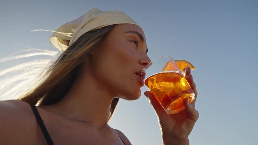 Closeup of a woman sipping a bright orange cocktail against a clear blue sky