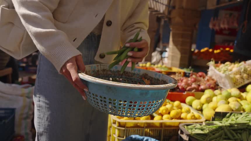 A woman carefully adds green beans to a blue basket shes holding with other fruit and produce visible in the background