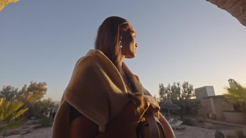 A young woman wearing a beige shawl and facing away from the camera is framed by a rough arched stone opening as she looks out over a sunlit arid landscape featuring palm trees and low buildings with warm light illuminating the scene