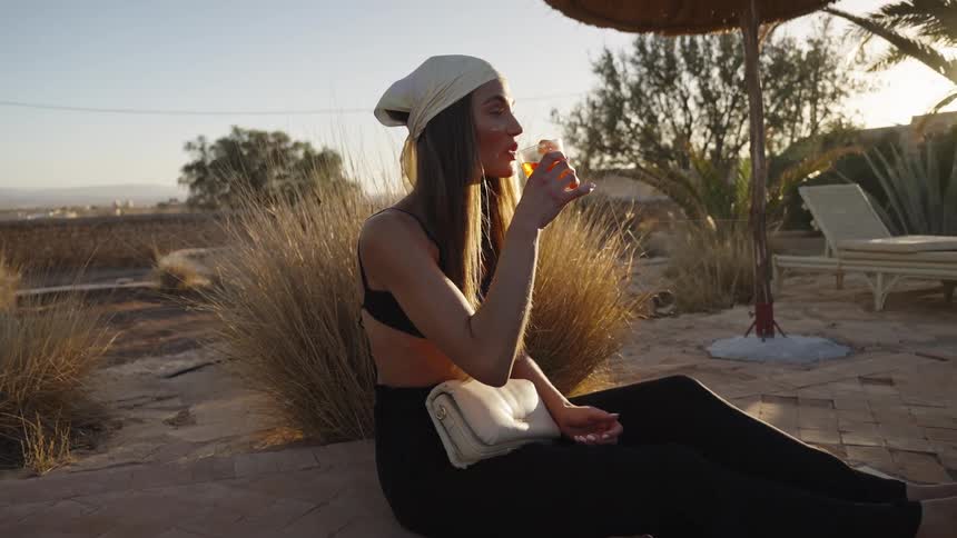 A young woman wearing a bandana and dark clothing sits on the terracotta tile edge of a pool holding and drinking a cocktail while enjoying the warm light and view of the arid outdoor setting