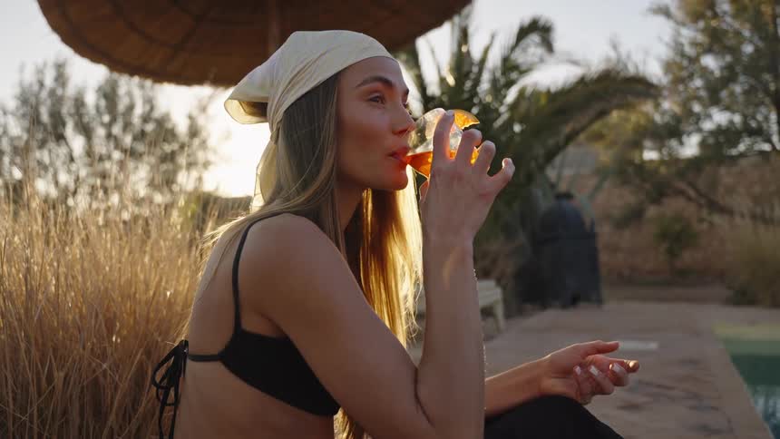 A longhaired woman wearing a black bikini top and a lightcolored bandana is seated next to a pool in a warm dry outdoor environment taking a sip from a glass containing an orangecolored cocktail