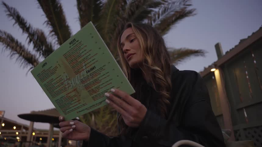 A woman reads a green menu at an outdoor restaurant at twilight