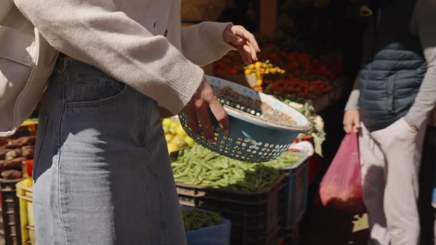 A closeup shot captures a person wearing light blue jeans and a white sweater holding an empty blue basket reaching into a crate to pick up fresh brown onions at an outdoor fruit and vegetable market