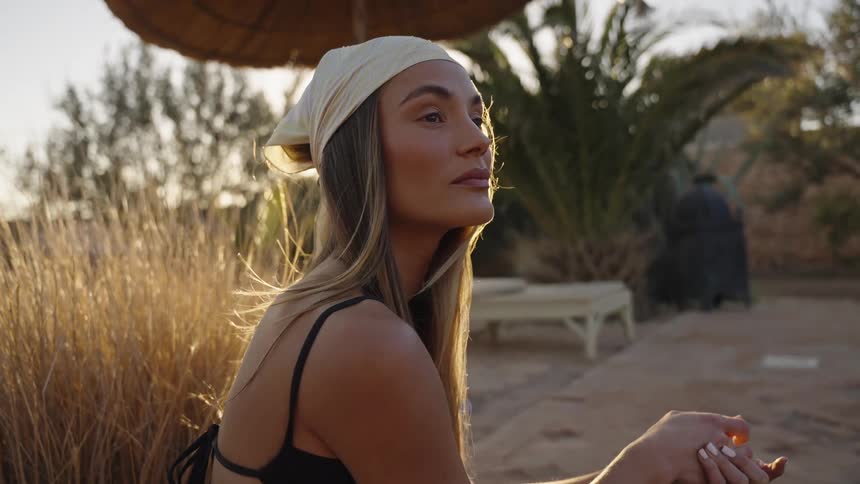 A young woman wearing a bandana and a dark top sitting outdoors in a sunlit dry landscape with palm trees as she looks offcamera with a thoughtful expression