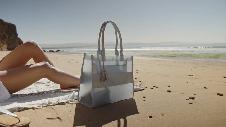 A light blue tote bag stands on a sunny beach next to the legs of a person relaxing on a towel by the water