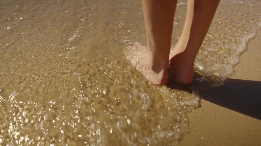 A closeup lowangle shot focuses on a persons bare feet standing in the goldenbrown sand as small gentle ocean waves wash over the toes and ankles