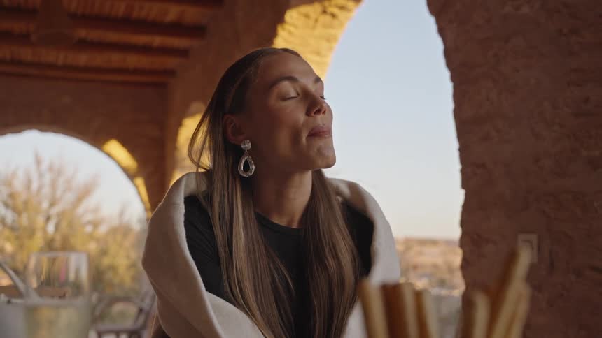A beautiful young woman with long dark hair and large earrings sits eating wrapped in a white shawl looking upwards in the bright light under a stone archway
