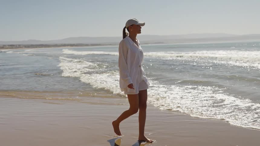 A woman in a white shirt and baseball cap walks barefoot along the wet sand of a sunny beach as small waves break near the shore