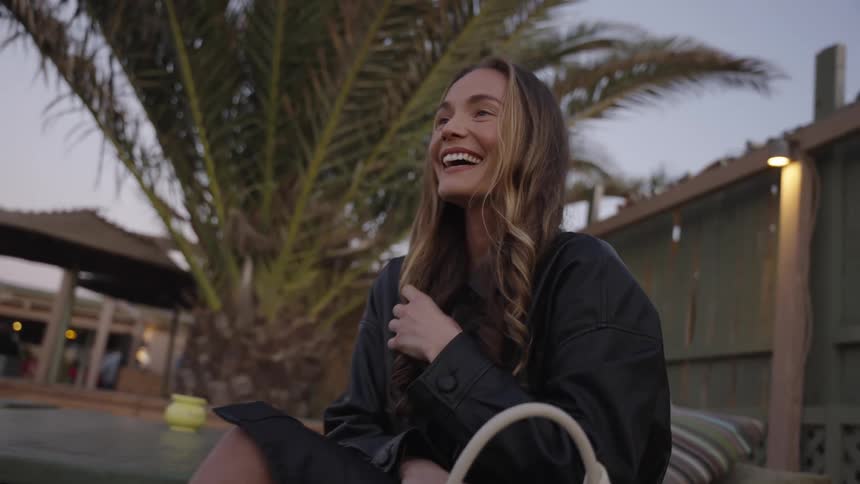 A beautiful young woman with long wavy hair sits outdoors near a palm tree wearing a dark jacket smiling widely and laughing