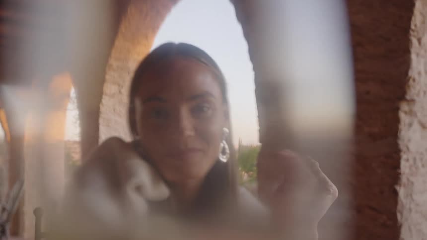 A smiling woman wearing a lightcolored shawl and large hoop earrings holds a tall champagne flute towards the camera in a closeup shot with a background of stone arches and an outdoor patio setting