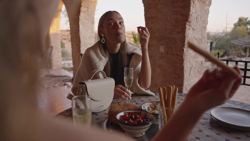 A beautiful woman sits at a table outdoors under a stone archway eating appetizers and drinking champagne with a friend in warm light
