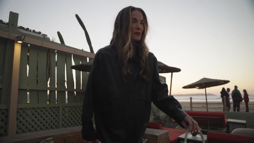 This video shows a woman dressed in a black coat or long shirt and carrying a lightcolored handbag walking onto the wooden patio of an outdoor lounge area she sits down on a cushioned bench facing the beach which is visible in the background with sun