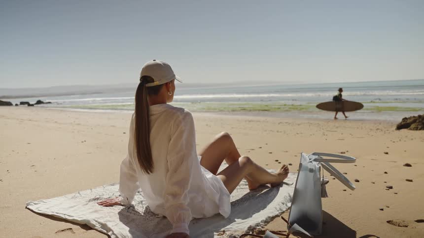 A woman sits on a sunny beach overlooking the ocean under bright sunlight