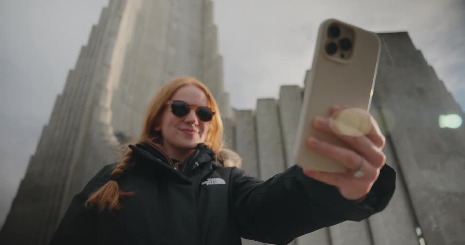 A close view shows a woman with red hair wearing sunglasses and a parka recording a selfie video in front of a massive gray vertical stone structure