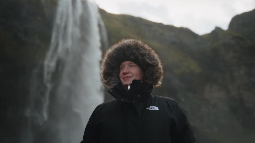 Woman in a black coat standing outdoors with a waterfall flowing in the distance