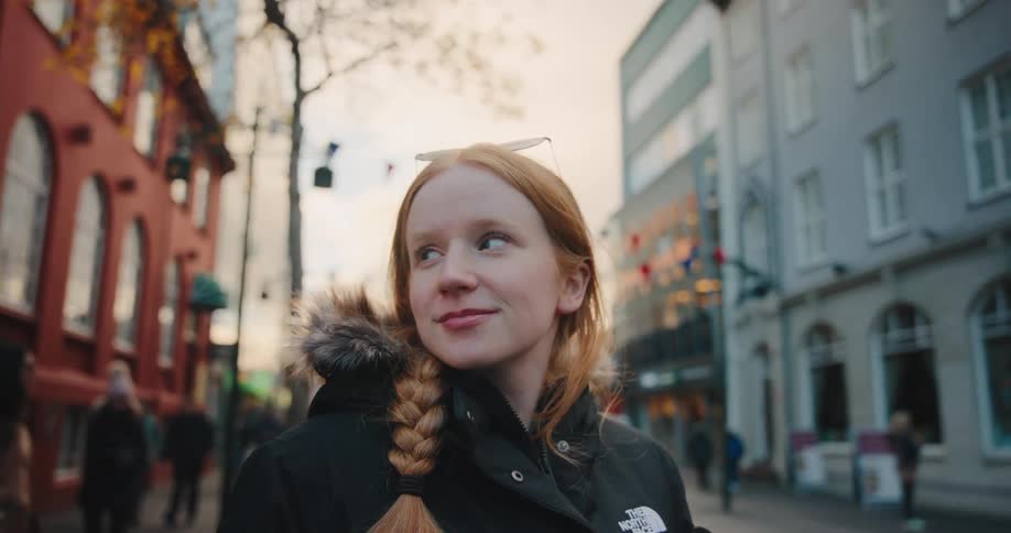 Person with braided red hair and black furlined jacket on sunlit urban street