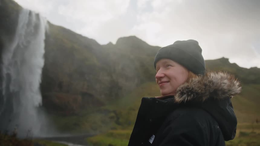 A young woman wearing a winter jacket and beanie smiles while looking toward a majestic waterfall dropping from rugged cliffs in a lush valley