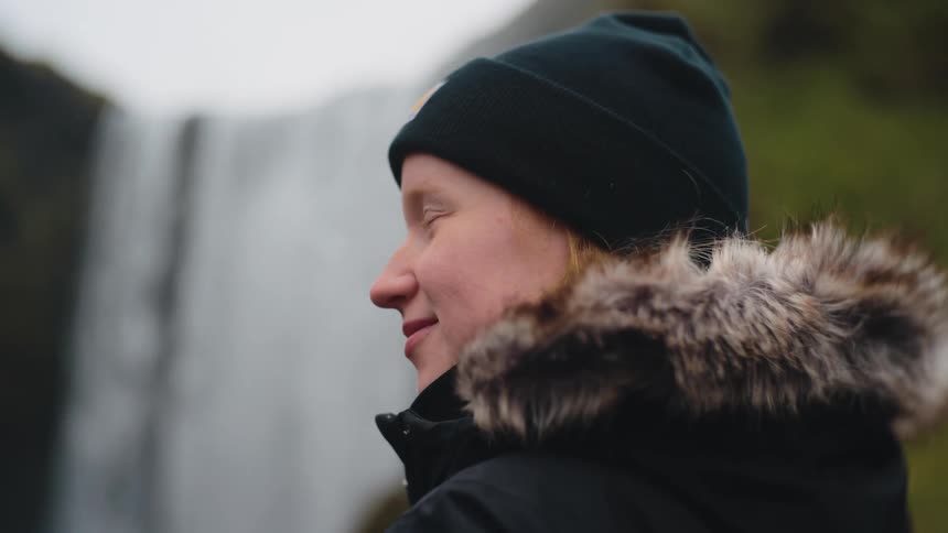 A smiling woman wearing a black beanie and winter coat stands in front of a blurred waterfall