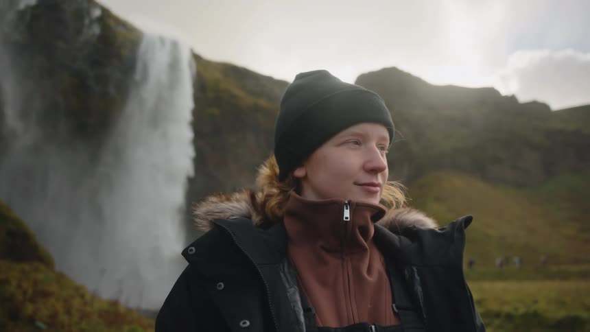 This video features a person with reddishblonde hair and a dark beanie wearing a heavy furtrimmed winter coat over a brown turtleneck layerthey are standing outdoors in a scenic landscape looking contemplative and smiling slightly with a large p