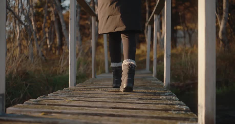 The camera follows the legs of a person wearing black leggings a dark coat and furlined boots as they walk across a rustic wooden footbridge the setting appears to be outdoors in a grassy area bordered by trees bathed in the soft warm light of late