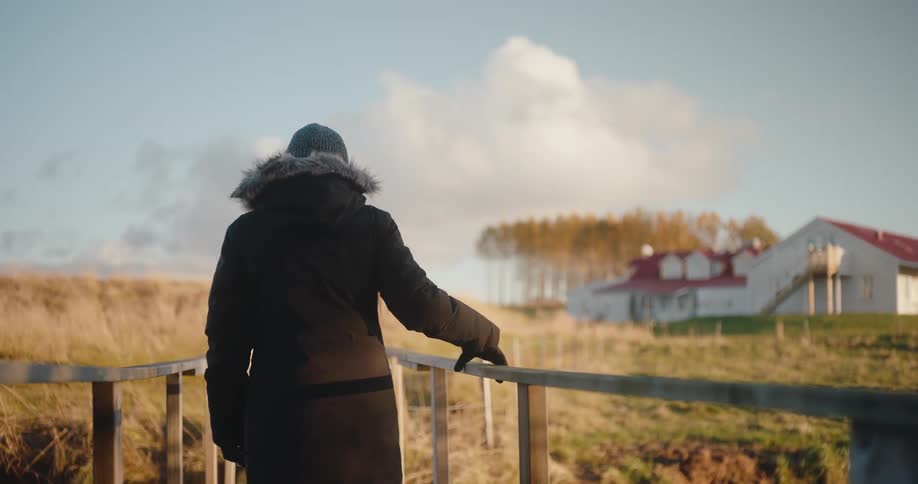A young woman in a winter jacket crosses a wooden bridge at midday heading toward a grassland with a building visible in the distance in the countryside