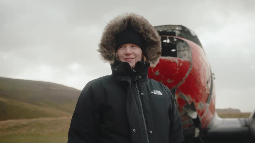 A smiling young woman in a fur trimmed parka and beanie stands in a stark landscape with the wreckage of a red and white airplane visible behind her