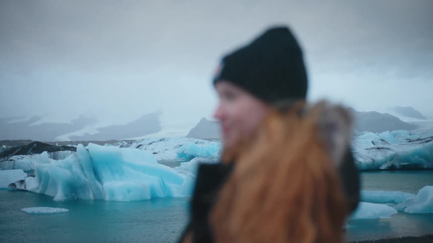 A blurred person with long hair and a beanie stands in the foreground overlooking a glacial lagoon filled with bright blue icebergs on a misty day