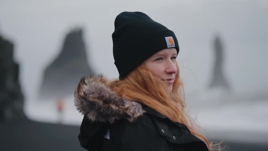 A young woman with long red hair and a black carhartt beanie is shown outdoors in a cold overcast environmentshe is wearing a dark winter coat with a furlined hood and in the foggy background several large rock formations or sea stacks are visible